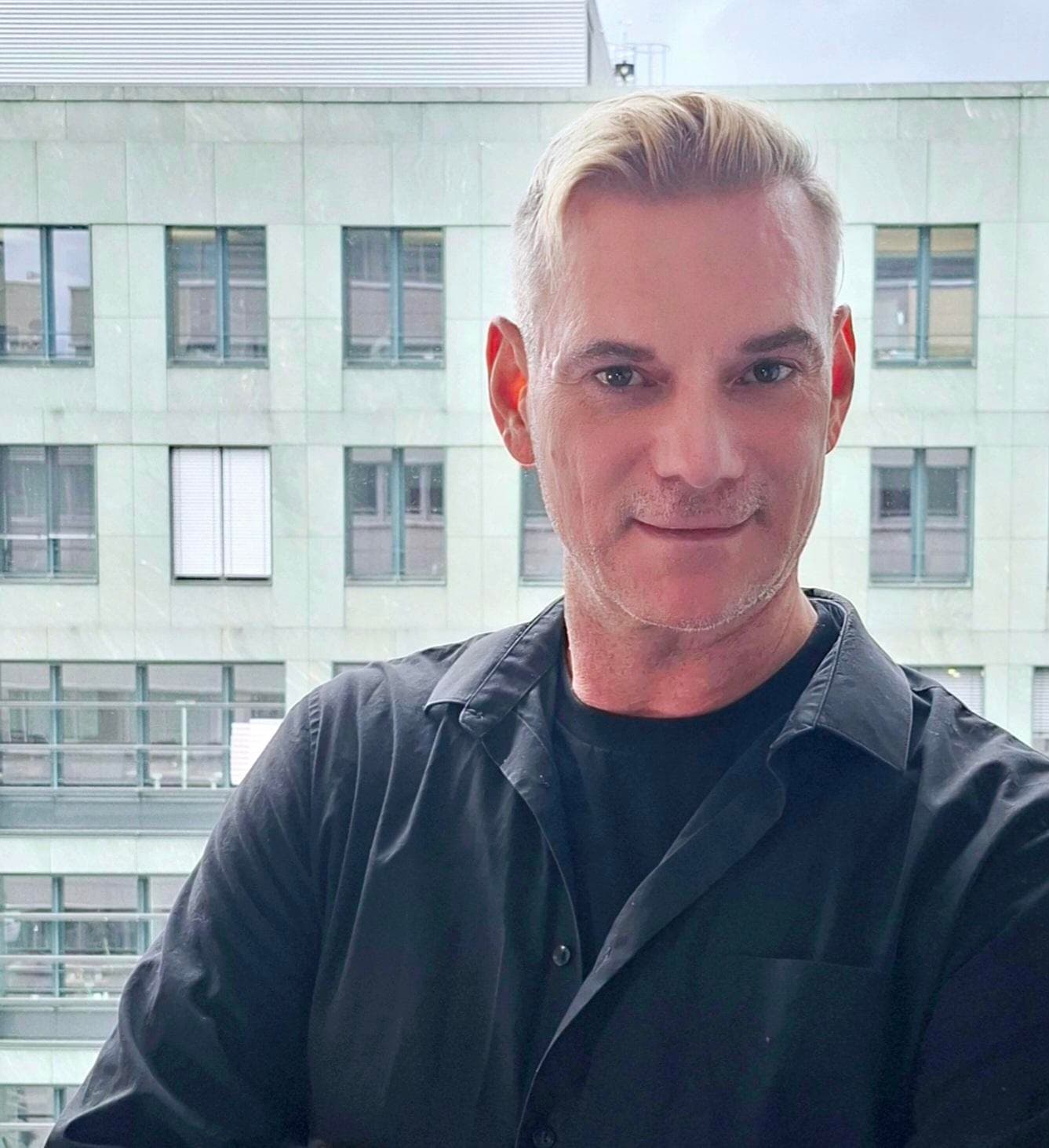 Mayk Int-Velt mit schwarzem Hemd vor einem Bürogebäude, blickt freundlich in die Kamera.