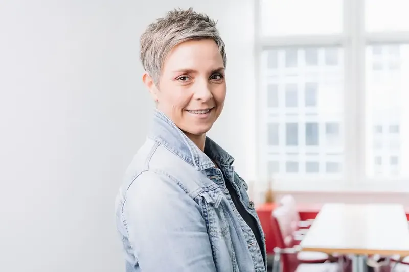 Kathrin mit kurzem Haar und Jeansjacke, lächelt freundlich im Büro.